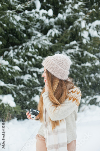 Beautiful young woman holding a retro vintage film camera for taking photos in winter snowy forest. Hobby and leisure activity outside. Hipster stylish girl in beanie hat in winter outdoors near fir.