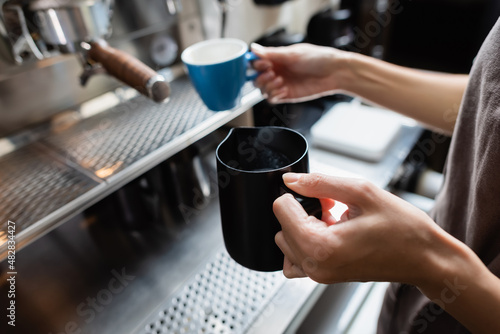 Cropped view of barista holding milk jug and blurred cup near coffee machine in cafe.