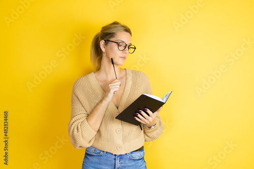 Young beautiful woman wearing casual sweater with a book in hand over isolated yellow background thinking and looking at the notebook