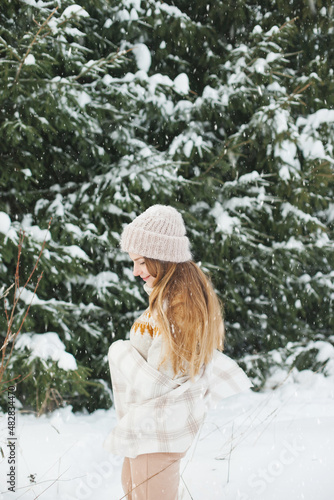 Happy woman in winter forest during snowfall, enjoying winter. Millennial young woman with long hair in beige warm outfit walking in the snowy park in nature near fir trees, being careless and free..