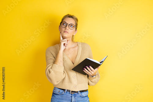 Young beautiful woman wearing casual sweater with a book in hand over isolated yellow background thinking, feeling doubtful and confused, with different options, wondering which decision to make.
