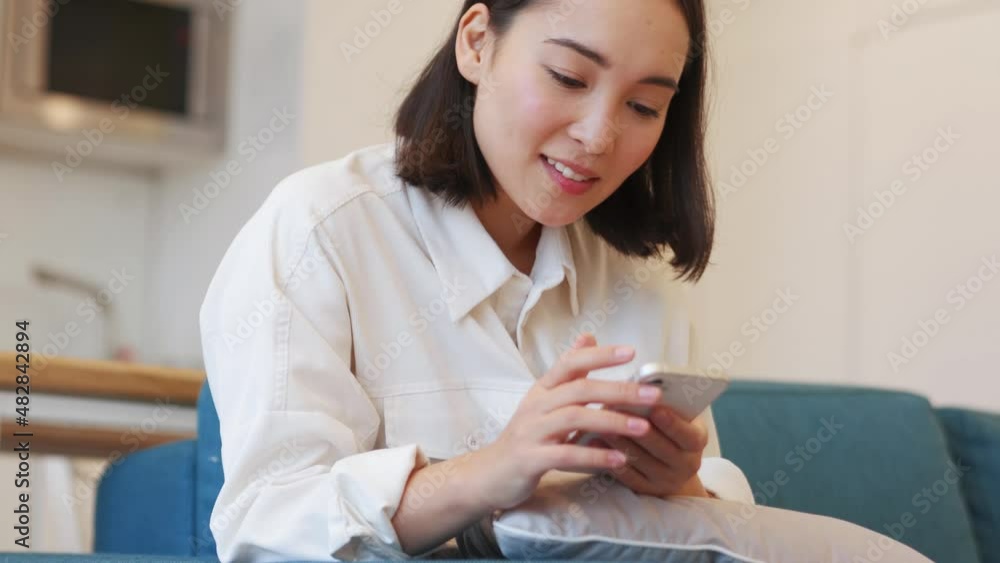 Positive Asian woman texting by phone on the sofa Positive Asian woman texting by phone on the sofa