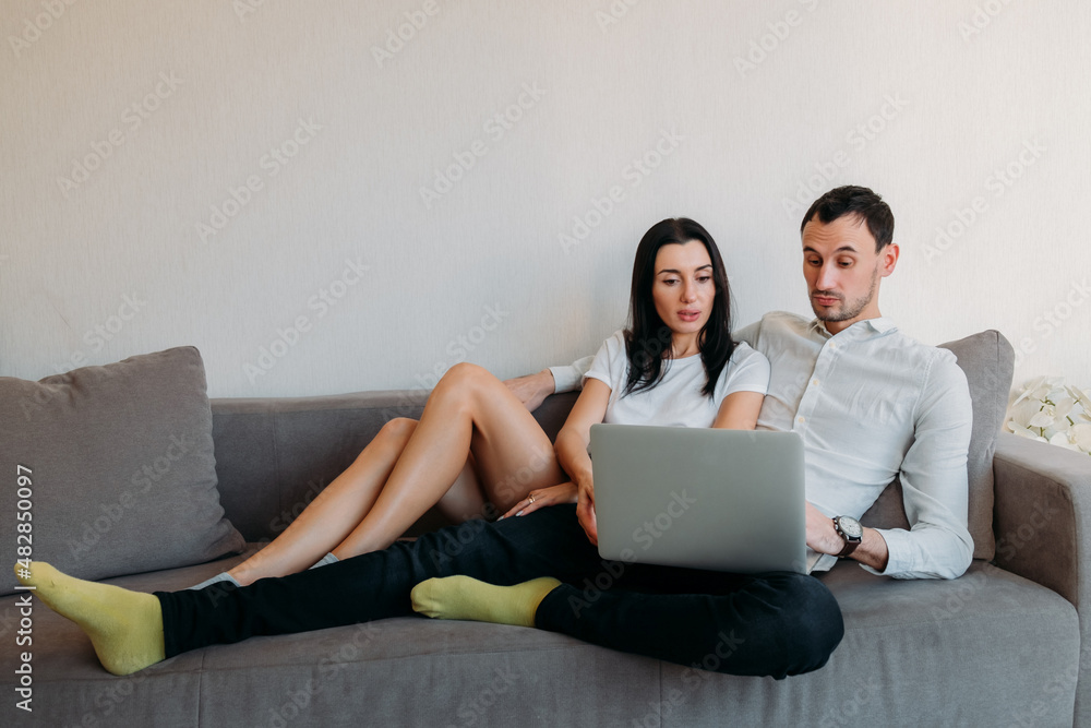 Young couple is lying on couch at home using laptop computer. Man looking at laptop screen is slightly surprised. Young couple is lying on couch at home using laptop computer. Man looking at laptop screen is slightly surprised.
