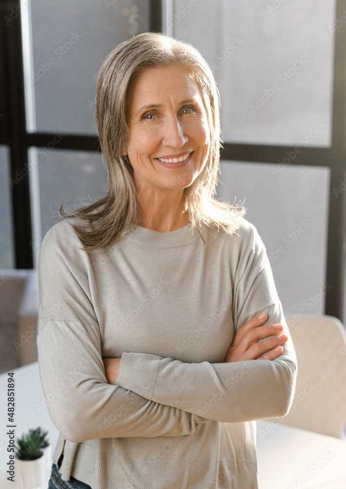 Fototapeta premium Vertical portrait of senior mature gray-haired female professional, leader, coach, elder businesswoman standing with her arms crossed folded in the office and confidently looking at the camera