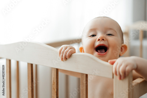 portrait of a 5 month old baby girl standing in a crib in a bright nursery after sleep and looking and smiling at the camera