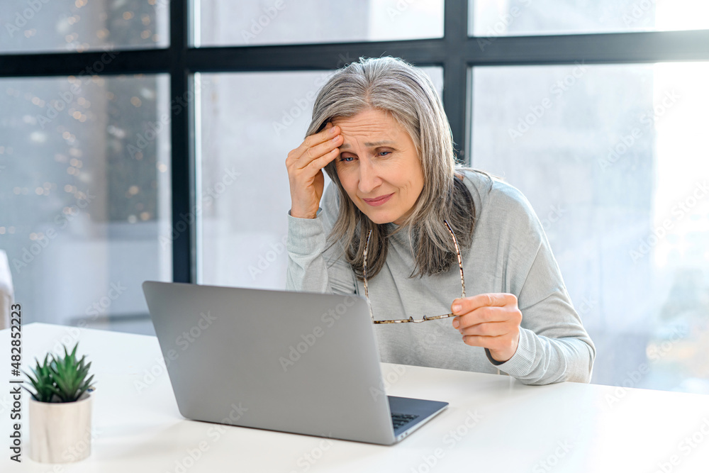 Concentrated upset senior woman sitting at the desk in front of laptop deep in thoughts and doubt, disappointed and worried female freelancer holding head with hands, has trouble and difficulties Concentrated upset senior woman sitting at the desk in front of laptop deep in thoughts and doubt, disappointed and worried female freelancer holding head with hands, has trouble and difficulties