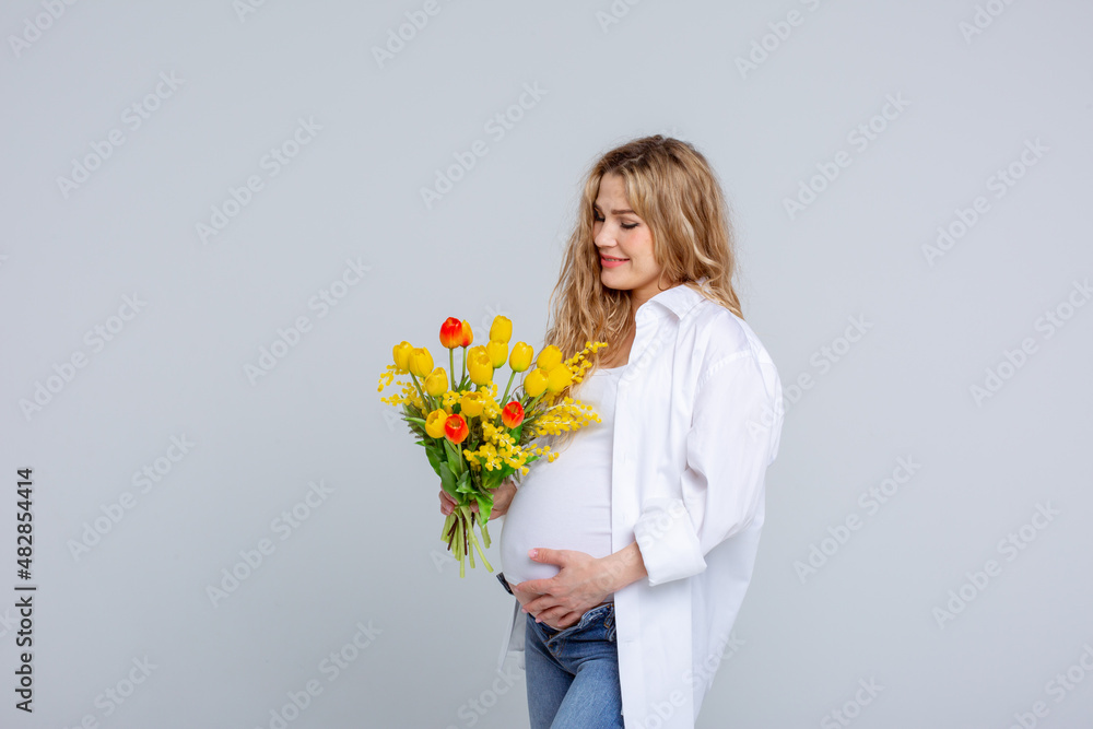 Concept Of A Happy Pregnancy. The Expectant Mother Touches Her Stomach holding a bouquet Of spring flowers Standing On A White Background. Studio shot Concept Of A Happy Pregnancy. The Expectant Mother Touches Her Stomach holding a bouquet Of spring flowers Standing On A White Background. Studio shot