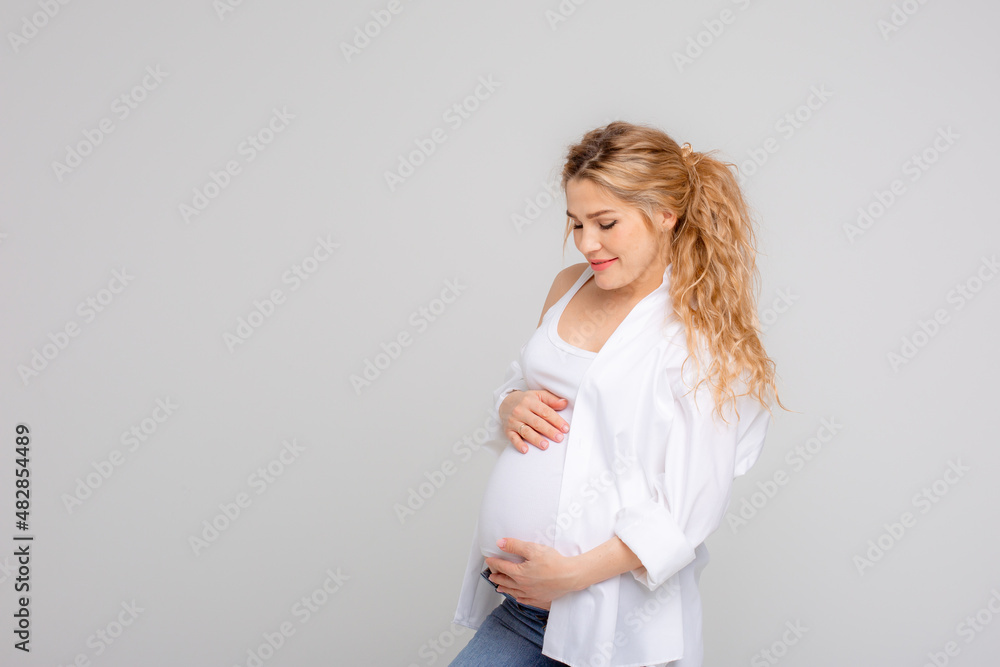 Concept Of A Happy Pregnancy. The Expectant Mother Touches Her Stomach, Smiling At The Camera Standing On A White Background. Studio shot Concept Of A Happy Pregnancy. The Expectant Mother Touches Her Stomach, Smiling At The Camera Standing On A White Background. Studio shot
