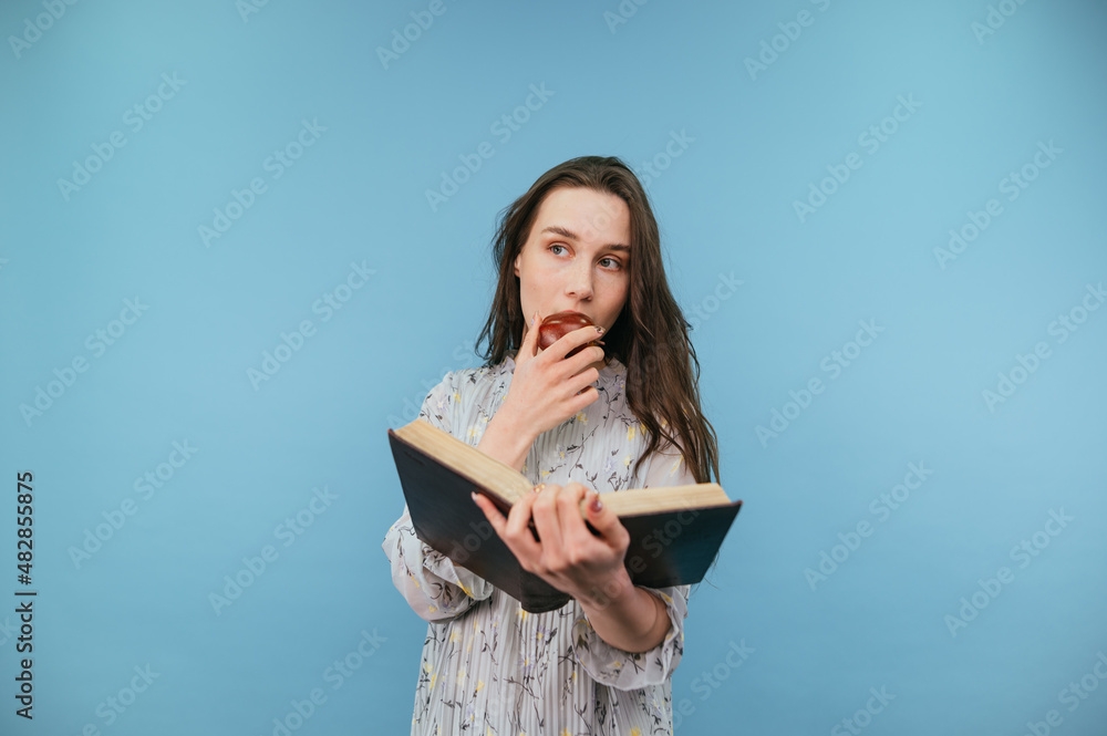 Beautiful woman in a dress with dark hair reads a book and eats an apple on a blue background, looking away with a serious face. Beautiful woman in a dress with dark hair reads a book and eats an apple on a blue background, looking away with a serious face.