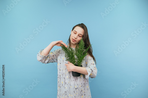 Attractive brunette in a dress isolated on a blue background with a flowerpot of ferns in her hands, stroking the plant and smiling.