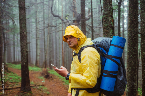 Male hiker in a yellow raincoat in the rain on a hike uses a smartphone against the backdrop of a foggy forest.