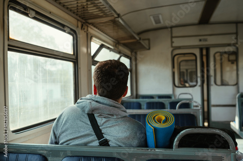 The back of a tourist man rides in an old train in the mountains with a backpack and sports mat.