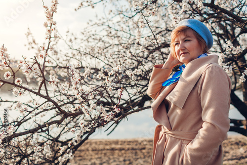 International Womens Day. Stylish senior woman walking in spring blooming park by river admiring nature landscape.