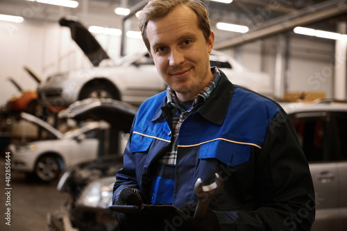 Handsome smiling Caucasian auto mechanic  technician in uniform  holding a wrench and standing against lifted cars with open hood in the repair shop