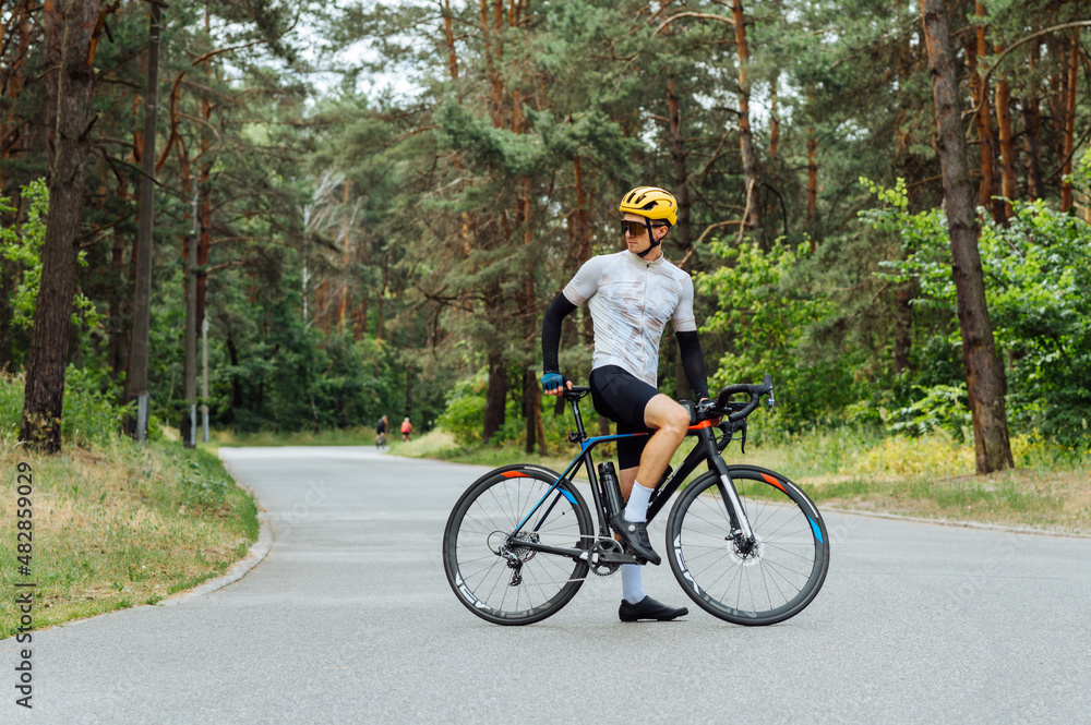 Athletic male cyclist in professional outfit stands with a bicycle on a forest road. Athletic male cyclist in professional outfit stands with a bicycle on a forest road.