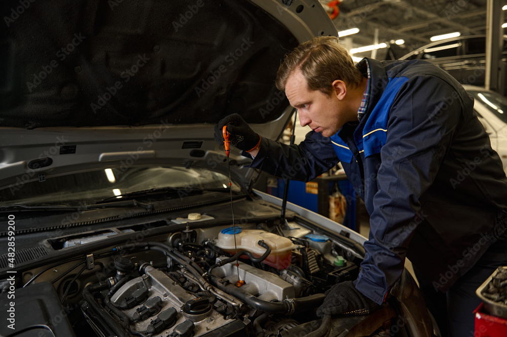 Concentrated Caucasian young man, car engineer technician mechanic in professional uniform, automobile mechanic repairman testing and checking oil level in the car engine. Car service and maintenance
