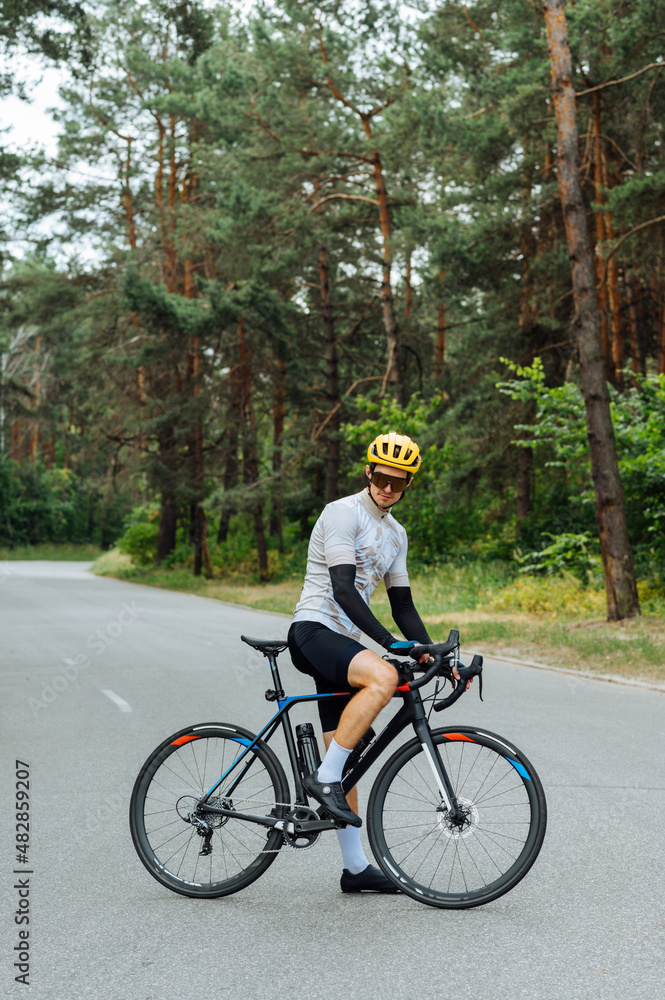 Athletic young man stands with a bicycle in the woods on the road, wears sports equipment and poses for the camera on the background of a beautiful view of nature.