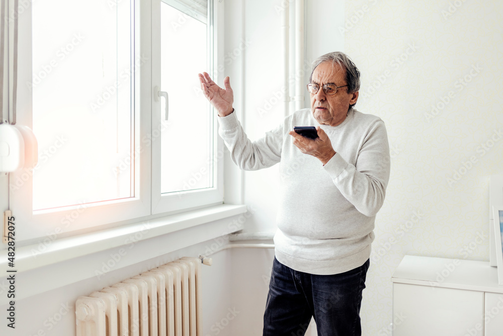 Fototapeta premium An unhappy elderly man standing next to a cold radiator, using a cell phone to call a technical service. Shot of an angry senior man talks to a technician about a radiator that doesn't work.