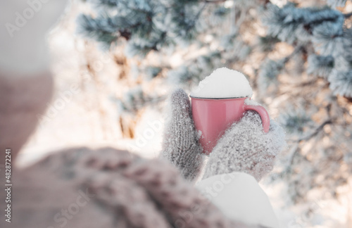 Winter image of woman's hands in the wool mittens holding vintage pink mug full of clear white snow. Winter freshness concept. Awesome snowy forest in sunny winter morning. Winter weekend out of town.