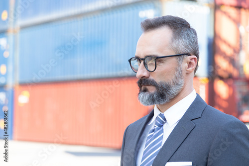 Portrait good looking Logistics manager wearing formal grey suite, white hard hat standing and visit his cargo warehouse and container boxes area.