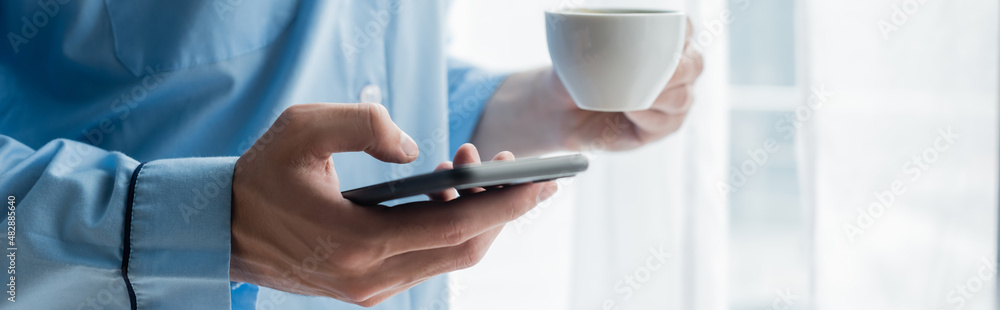 cropped view of man in blue pajamas using cellphone while holding coffee cup, banner. cropped view of man in blue pajamas using cellphone while holding coffee cup, banner.