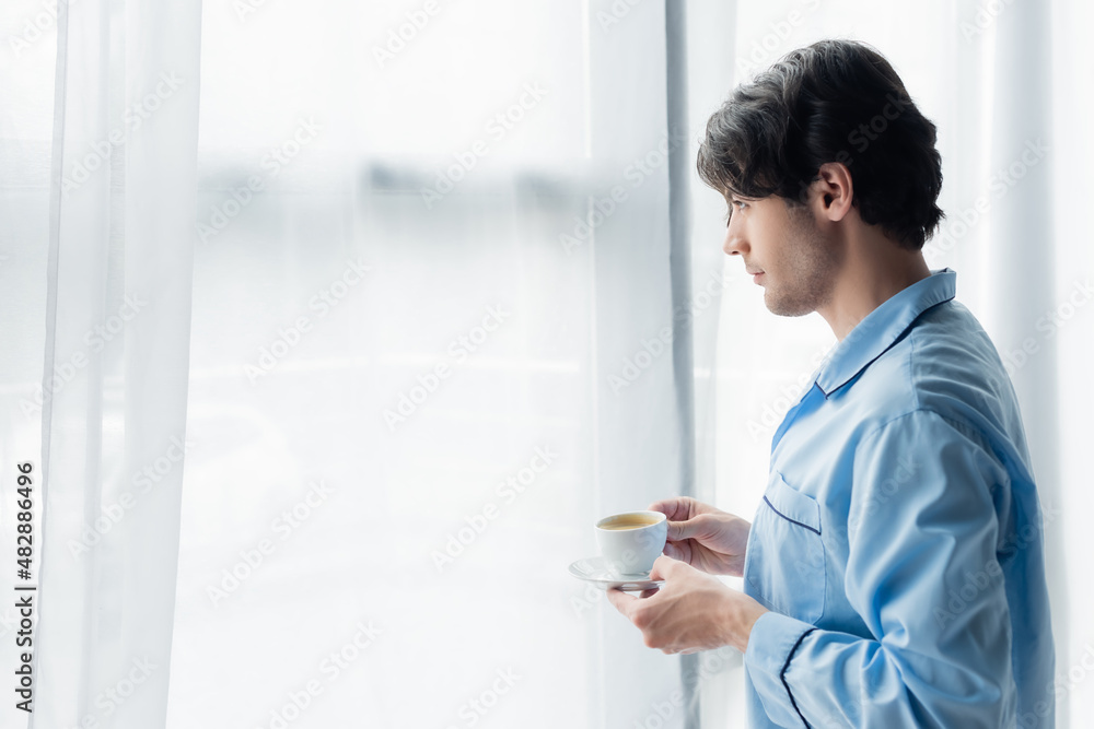 side view of brunette man in blue pajamas standing with cup of coffee near window. side view of brunette man in blue pajamas standing with cup of coffee near window.