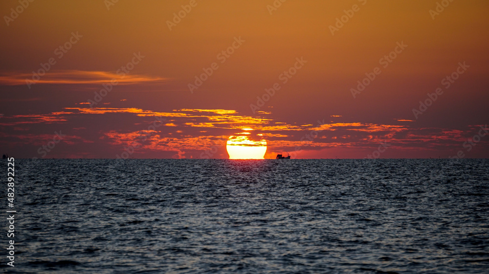 Ship floating on the water towards the horizon in the rays of the setting sun. Beautiful orange sky and clouds with yellow reflections