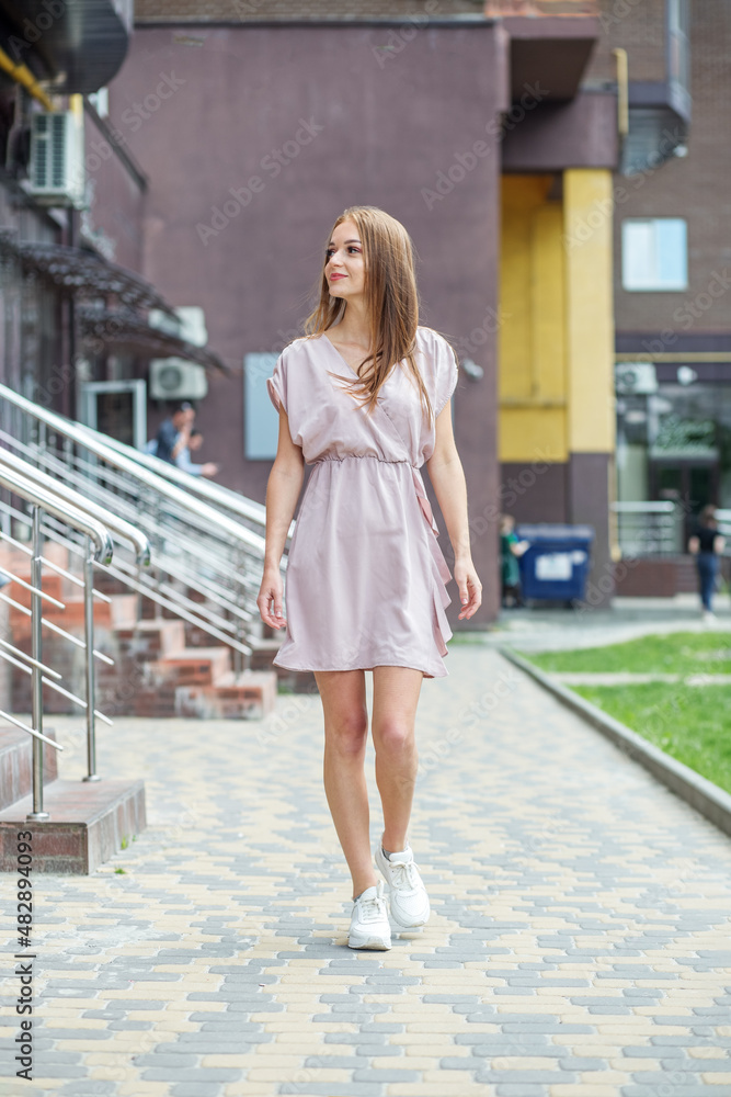 Young happy woman walks around city on summer day. Dressed in light dress and white sneakers.