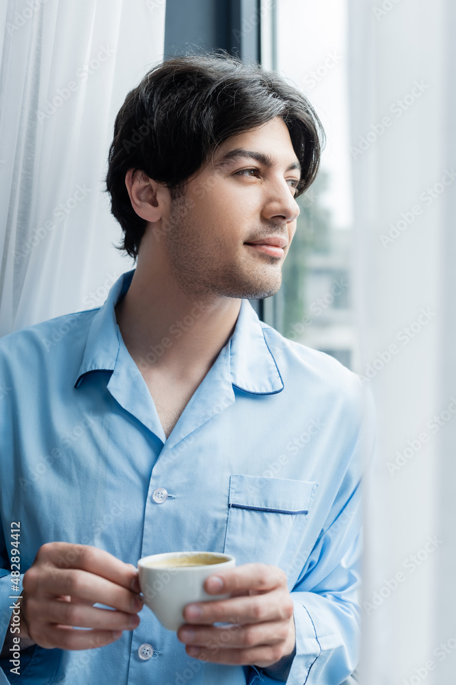 young brunette man in blue pajamas standing with coffee cup near window. young brunette man in blue pajamas standing with coffee cup near window.
