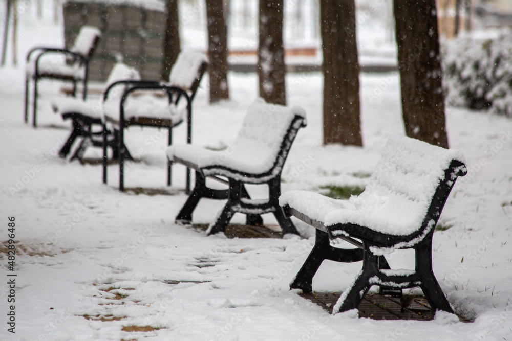 An empty bench in a garden while it's snowing. Bench covered with snow