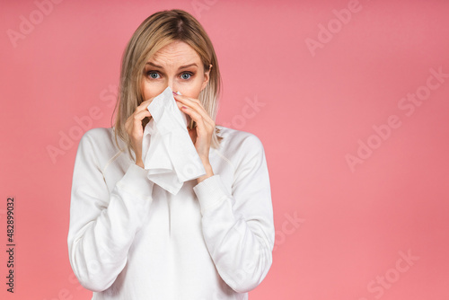 Studio portrait of an unhealthy pretty blonde woman in casual blowing her nose napkins, looking at the source of the allergy, rhinitis, cold, allergy concept isolated over pink background.