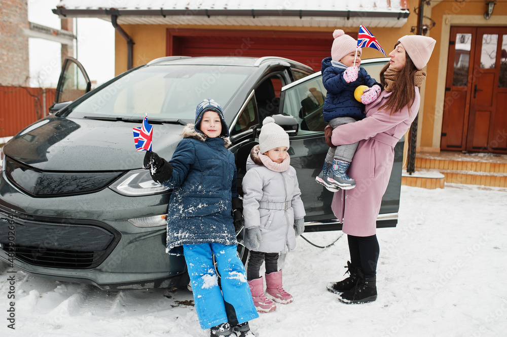 Young british mother with kids hold Great Britain flags and charging electric car in the yard of her house at winter.