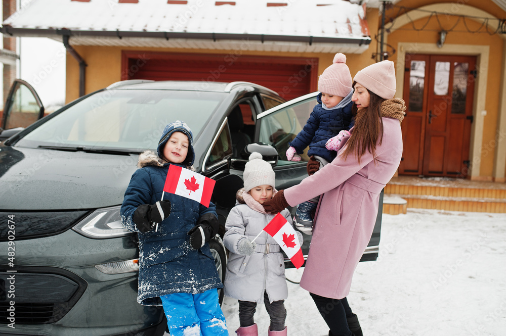 Young canadian mother with kids hold Canada flags and charging electric car in the yard of her house at winter. Young canadian mother with kids hold Canada flags and charging electric car in the yard of her house at winter.