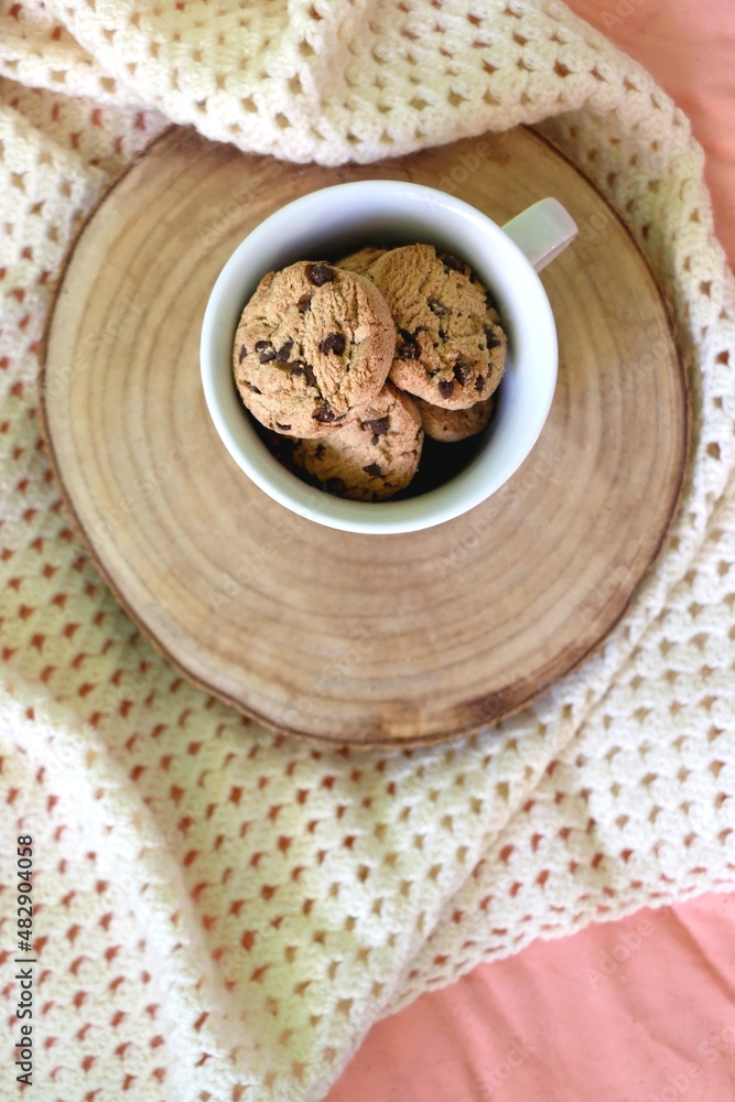 Fototapeta premium Mug filled with chocolate chip cookies and soft knitted blanket on a bed. Flat lay.