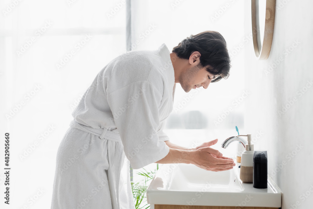 side view of young man in white bathrobe near sink and toiletries in bathroom.