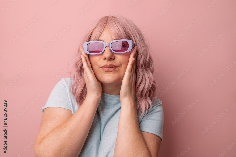 Fototapeta premium Happy young caucasian woman in bright stylish sunglasses and a blue t-shirt, smiling posing looking at the camera isolated over pink background.