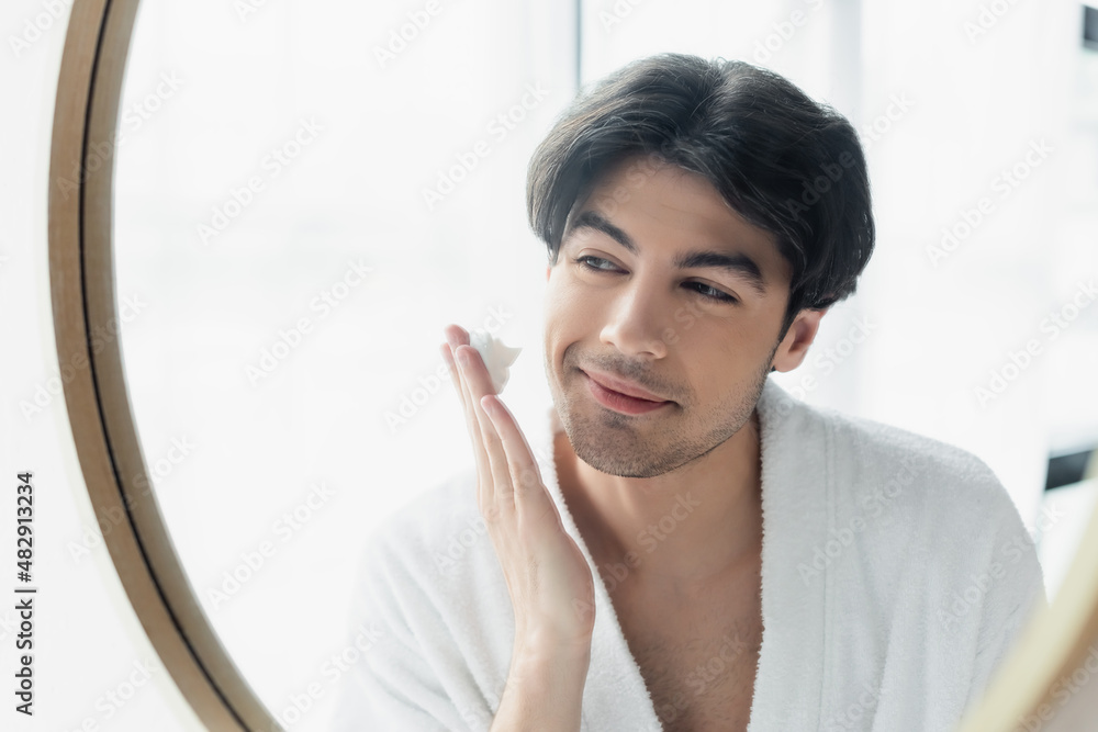 young smiling man in bathrobe applying shaving foam near mirror.