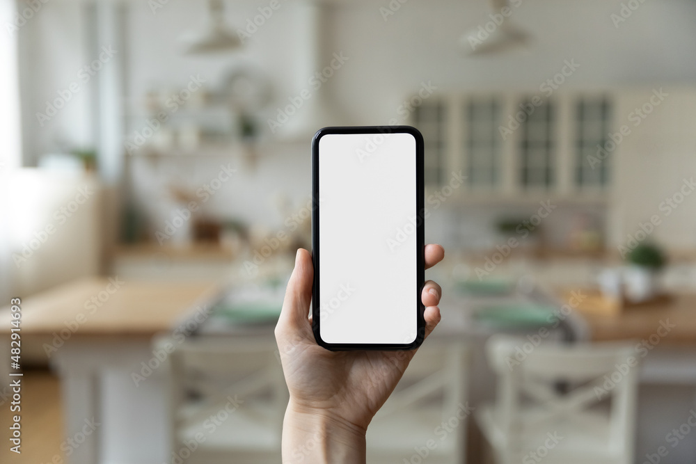Close up woman hand holds modern smartphone with white mock up blank screen, kitchen on blurred background. New mobile app software design, shopping on-line, web surfing, internet, connection concept