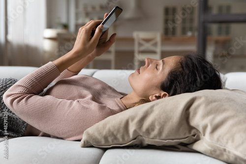 Serene woman relaxing lying on cushion on sofa with smartphone, reading media news, chatting online, make food delivery order through website retail services. Internet connection, modern tech concept