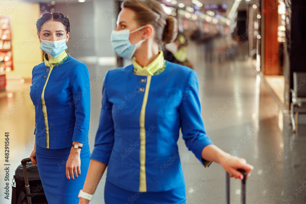 Women stewardesses wearing protective face masks and air hostess uniform while walking down airport terminal during pandemic