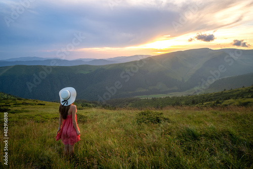 Young woman in red dress standing on grassy field on a windy evening in autumn mountains enjoying view of nature