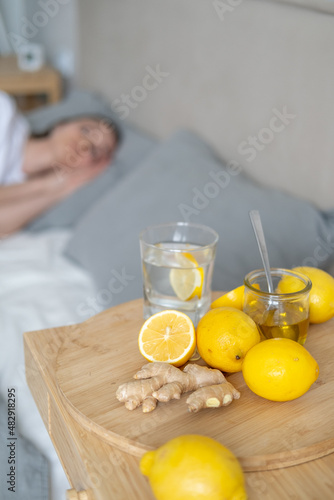 Sick girl in bed in the background. A glass of water on a wooden serving tray with ginger root, honey and a slice of lemon next to the bed in the bedroom