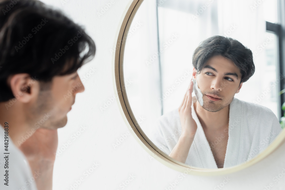 brunette man looking in bathroom mirror while applying shaving foam. brunette man looking in bathroom mirror while applying shaving foam.