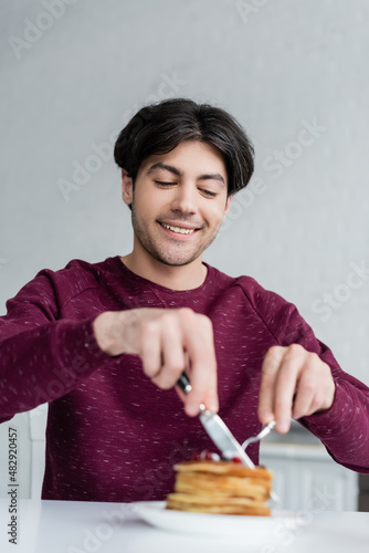 cheerful man cutting pancakes while having breakfast on blurred foreground.