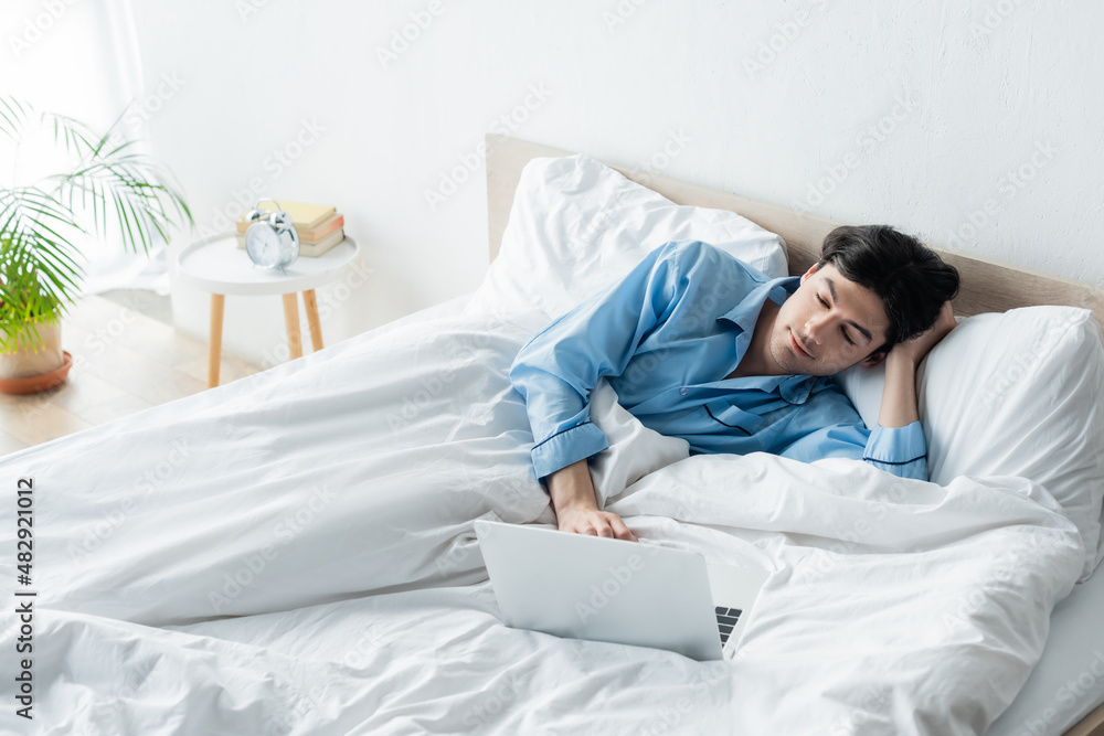 positive man in blue pajamas watching movie on laptop in bed. positive man in blue pajamas watching movie on laptop in bed.