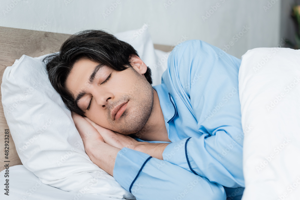 young brunette man in blue pajamas sleeping on white bedding. young brunette man in blue pajamas sleeping on white bedding.