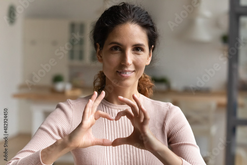 Head shot portrait beautiful loving young Latina woman makes heart symbol sign with joined fingers smile look at camera pose alone at home. Express gratitude, appreciation. I love you gesture concept photo