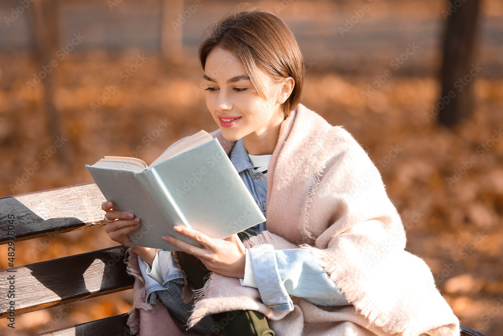 Obraz premium Young woman with plaid reading book on wooden bench in autumn park