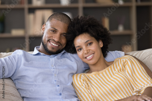 Beautiful young 35s African good-looking couple in love relaxing sitting on cozy sofa smiling looking at camera. Close up of happy homeowners family portrait. Romantic relationships, dating concept