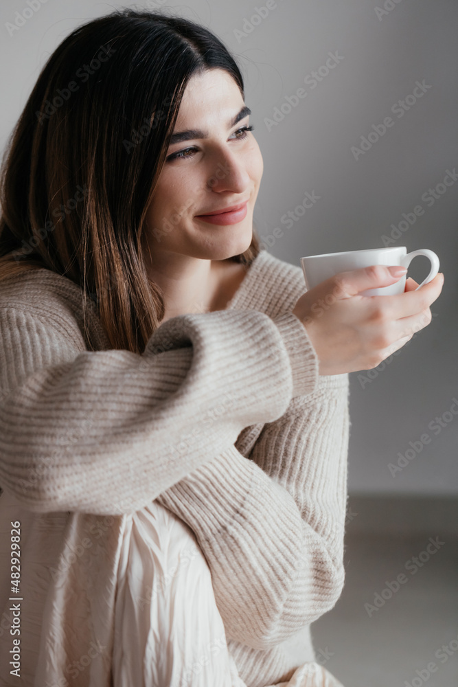 Beautiful young girl with brown hair in beige sweater looks away and holds a white cup in her hand. Portrait of girl smiling at home. Woman in warm sweater with cup of hot drink looking away Beautiful young girl with brown hair in beige sweater looks away and holds a white cup in her hand. Portrait of girl smiling at home. Woman in warm sweater with cup of hot drink looking away